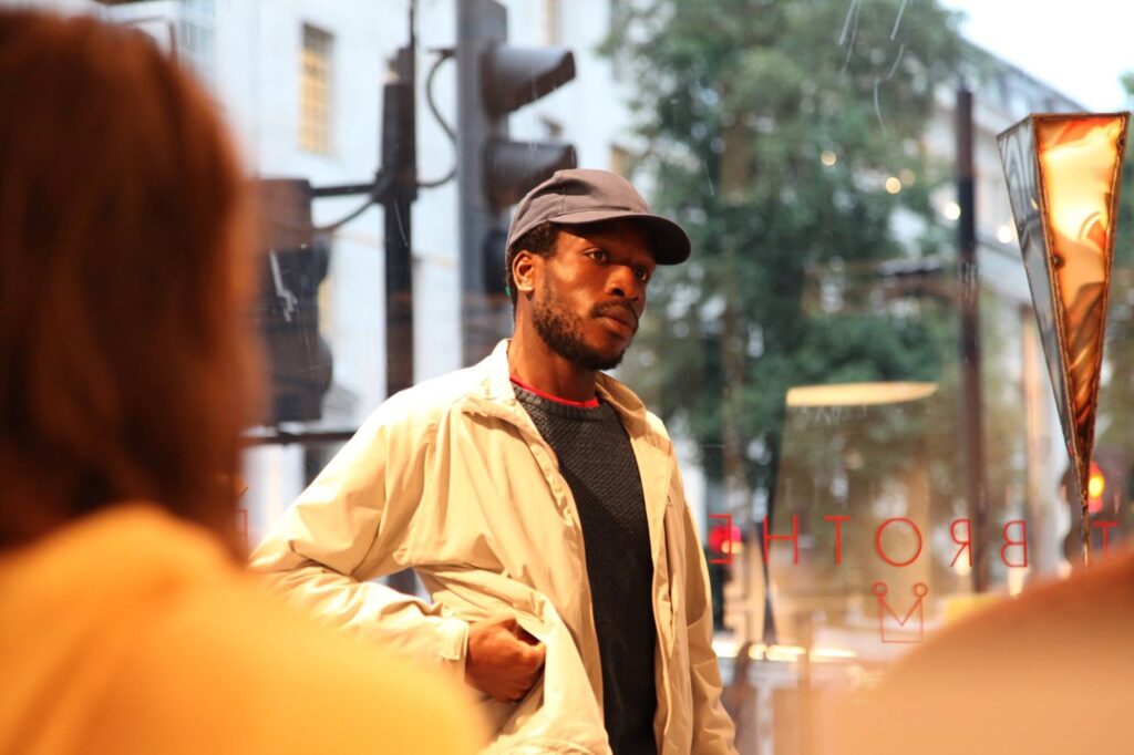 Black man in blue baseball cap, white jacket and blue jumper. Behind him is a large glass window at street level, outside we can see trees and traffic lights